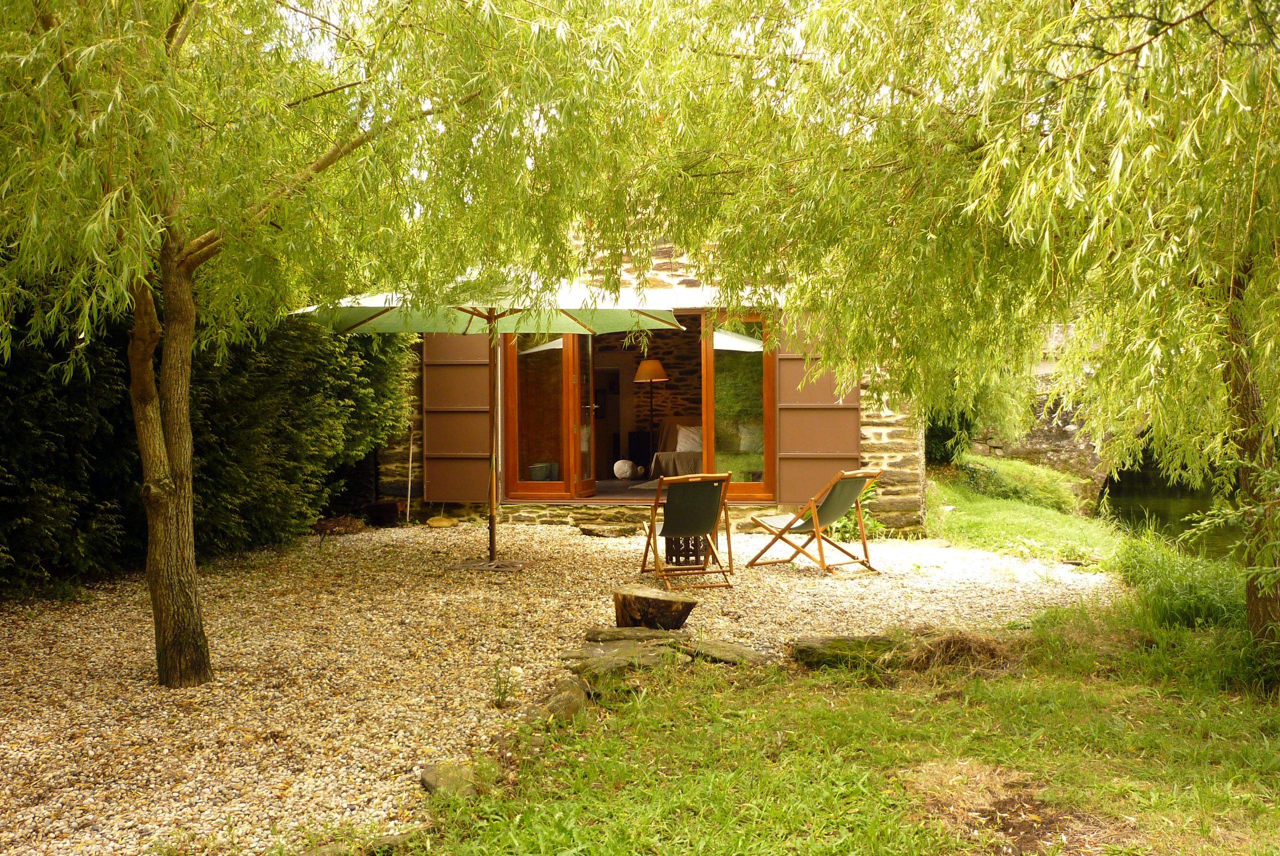 Picture contains a view of a small hut surrounded by trees and two outdoor chairs in the foreground. 